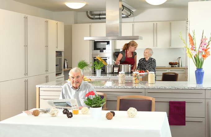 Senior sitzt an einem schön gedeckten Tisch, im Hintergrund die Wohnküche mit zwei Personen