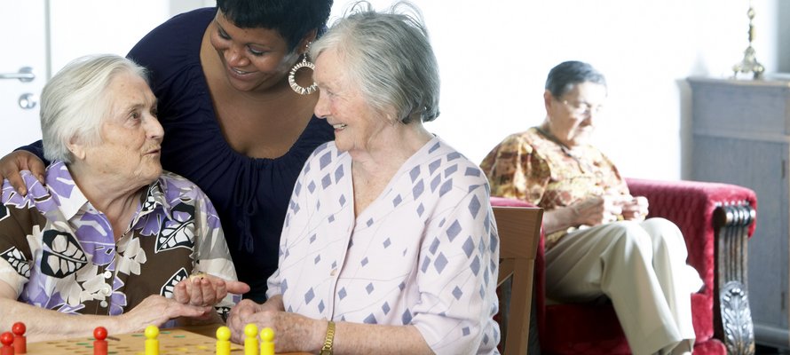 Betreuungskraft spricht mit zwei Seniorinnen, ein Senior sitzt im Hintergrund